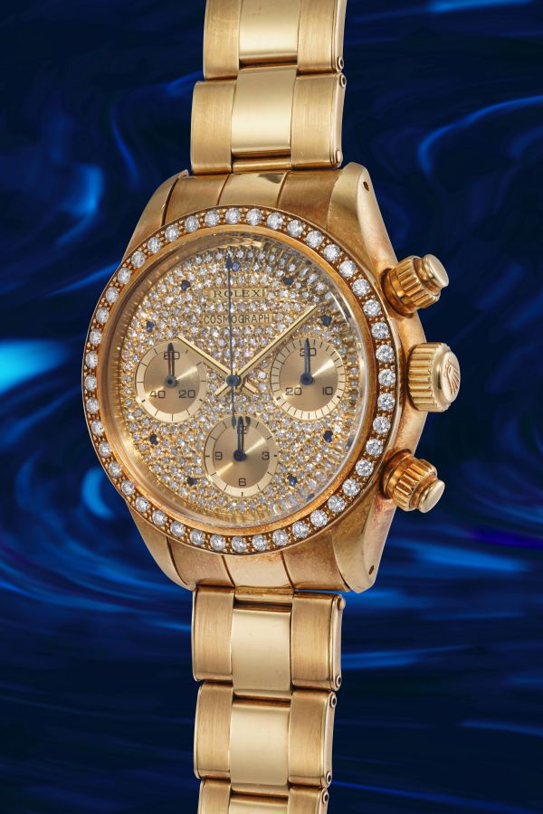 Un lussuoso orologio da polso in oro con quadrante e lunetta tempestati di diamanti, con tre contatori, si staglia su uno sfondo blu scuro e vorticoso.