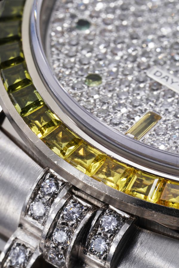 Primo piano del quadrante di un orologio di lusso tempestato di diamanti, con lunetta incastonata di pietre gialle e bracciale in metallo argentato anch'esso ornato di diamanti. Sul quadrante è parzialmente visibile la scritta "DAY".