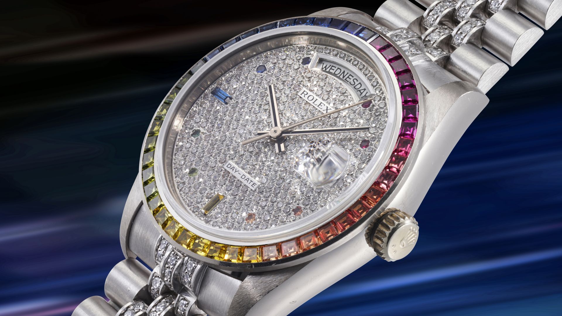 Un lussuoso orologio da polso Rolex Sultano con quadrante tempestato di diamanti, lunetta con gemme color arcobaleno e cinturino in metallo argentato, visualizzato su uno sfondo sfocato e multicolore.