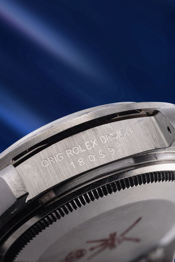 Primo piano della cassa di un orologio Rolex in acciaio inossidabile, che mostra il testo inciso "ORIG ROLEX DESIGN 18059" su uno sfondo blu sfocato.