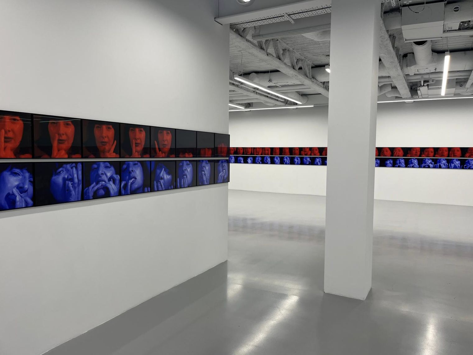 Una galleria d'arte moderna con pannelli fotografici su pareti bianche, che espongono ritratti ravvicinati di Marina Abramović di ispirazione londinese, dai toni blu e rossi, di volti che esprimono varie emozioni. Lo spazio ha un design minimalista e industriale.