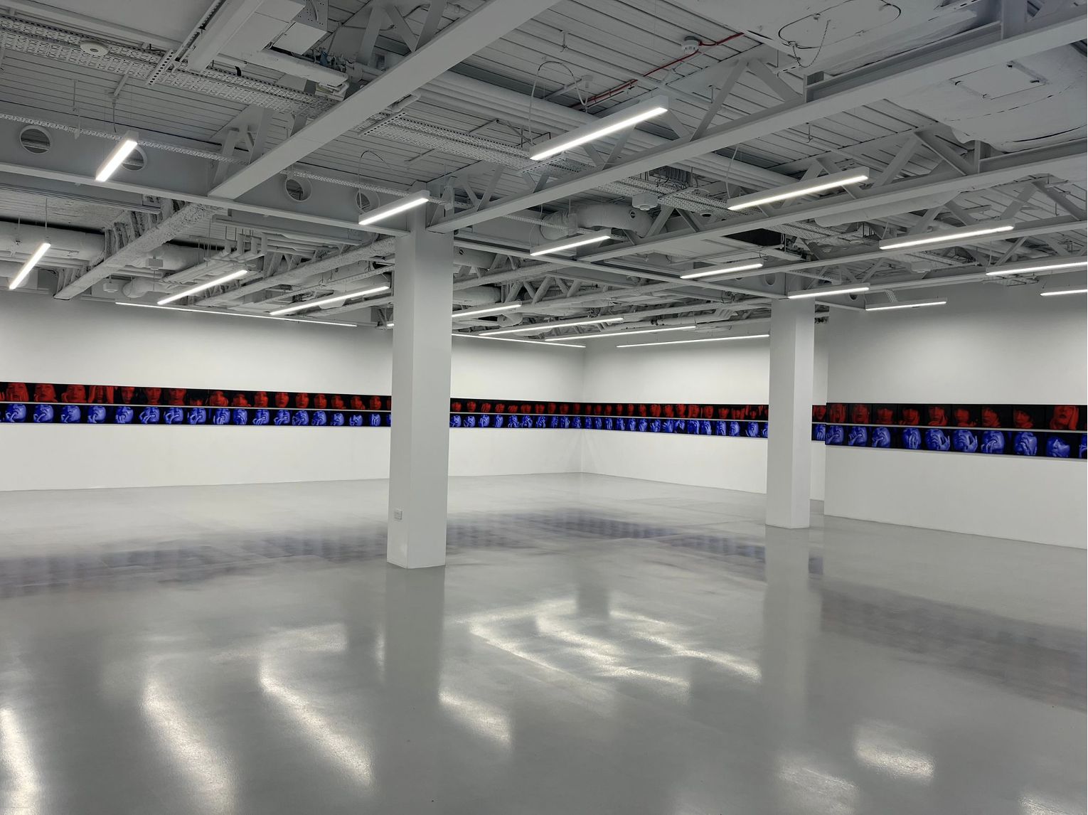 Una moderna e spaziosa galleria d'arte di Londra, con pavimenti lucidi e pareti bianche, espone una lunga striscia orizzontale di immagini colorate e astratte che ricordano Marina Abramović, che avvolge due lati della stanza sotto le luci del soffitto.