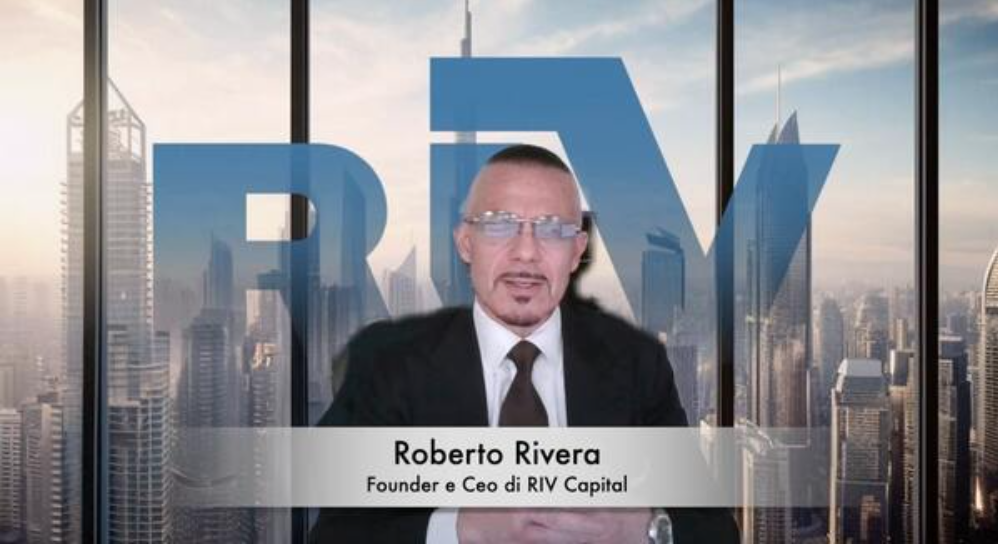 Un uomo in giacca e cravatta siede a una scrivania davanti a grandi finestre che inquadrano lo skyline della città. Alle sue spalle campeggia la scritta "Roberto Rivera, Founder e CEO di RIV Capital" e le lettere in grassetto "RIV".