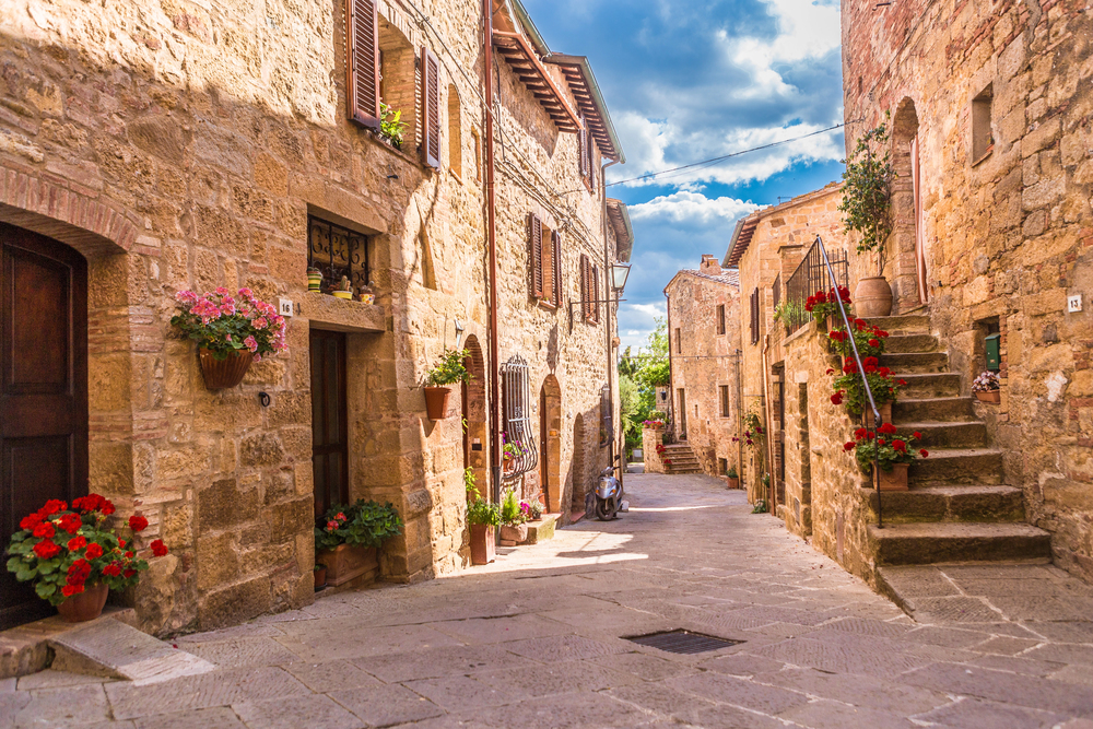 A narrow, sunlit street in a rustic village with stone buildings, wooden shutters, stairs, and many colorful flower pots lining the walls. The sky is bright blue with a few clouds.