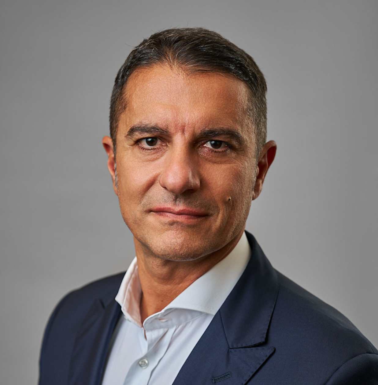 Un uomo con i capelli corti e scuri che indossa una giacca da abito blu e una camicia bianca con colletto posa per un'immagine professionale su uno sfondo grigio semplice, che rappresenta l'impegno di Weltix per le infrastrutture fintech innovative e i beni privati.