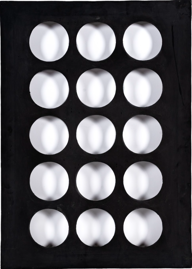 Un pannello nero rettangolare con una griglia di 15 ritagli circolari uniformemente distanziati evoca l'arte kruso. Lo sfondo bianco e le ombre creano un effetto tridimensionale e scultoreo che ricorda lo stile di Boetti.