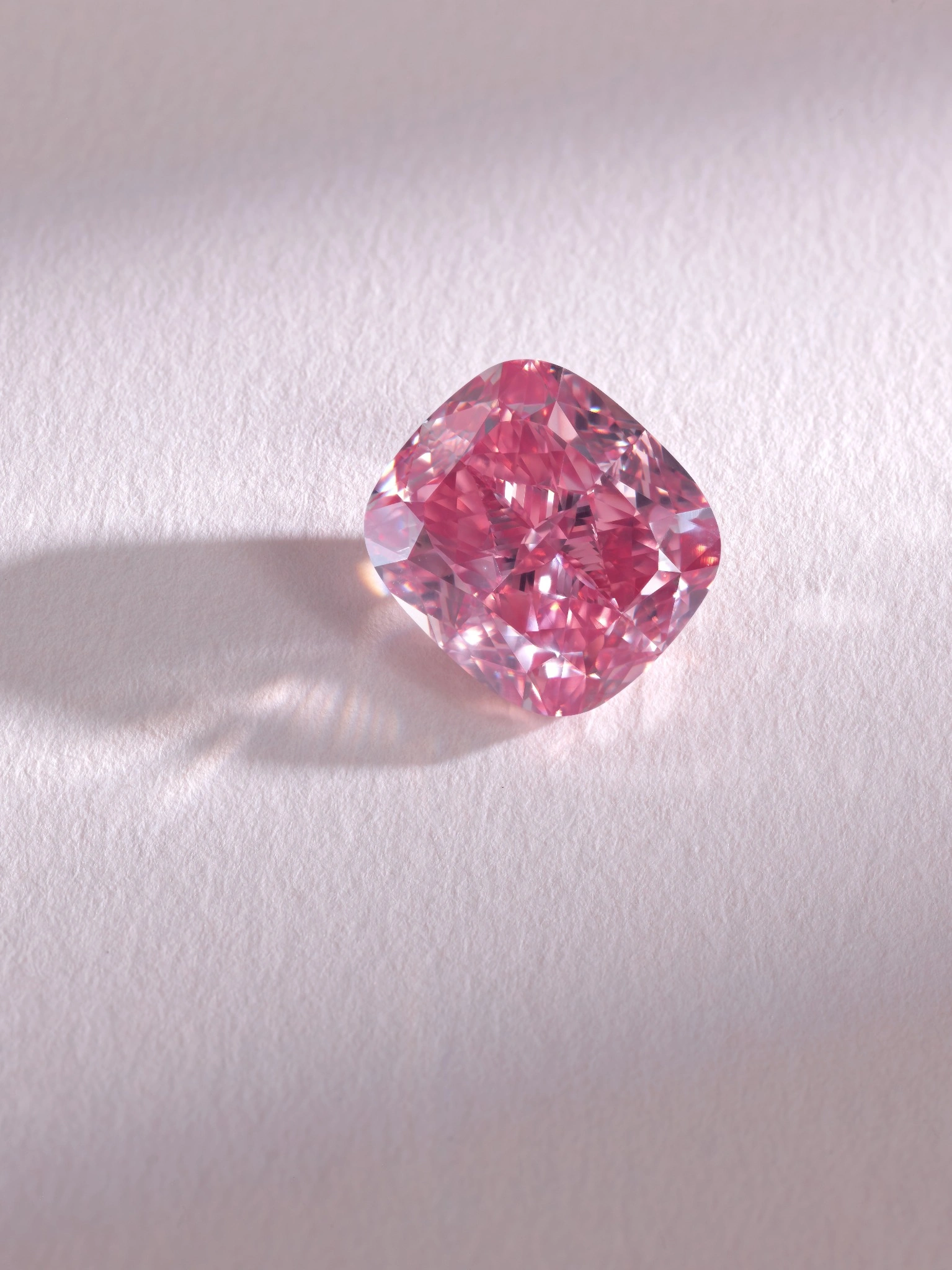 Un diamante rosa sotheby's con taglio a cuscino poggia su una superficie chiara e strutturata, proiettando un'ombra morbida a sinistra. Le sfaccettature del diamante scintillano, mostrando vibranti sfumature rosa e viola sotto una delicata illuminazione.