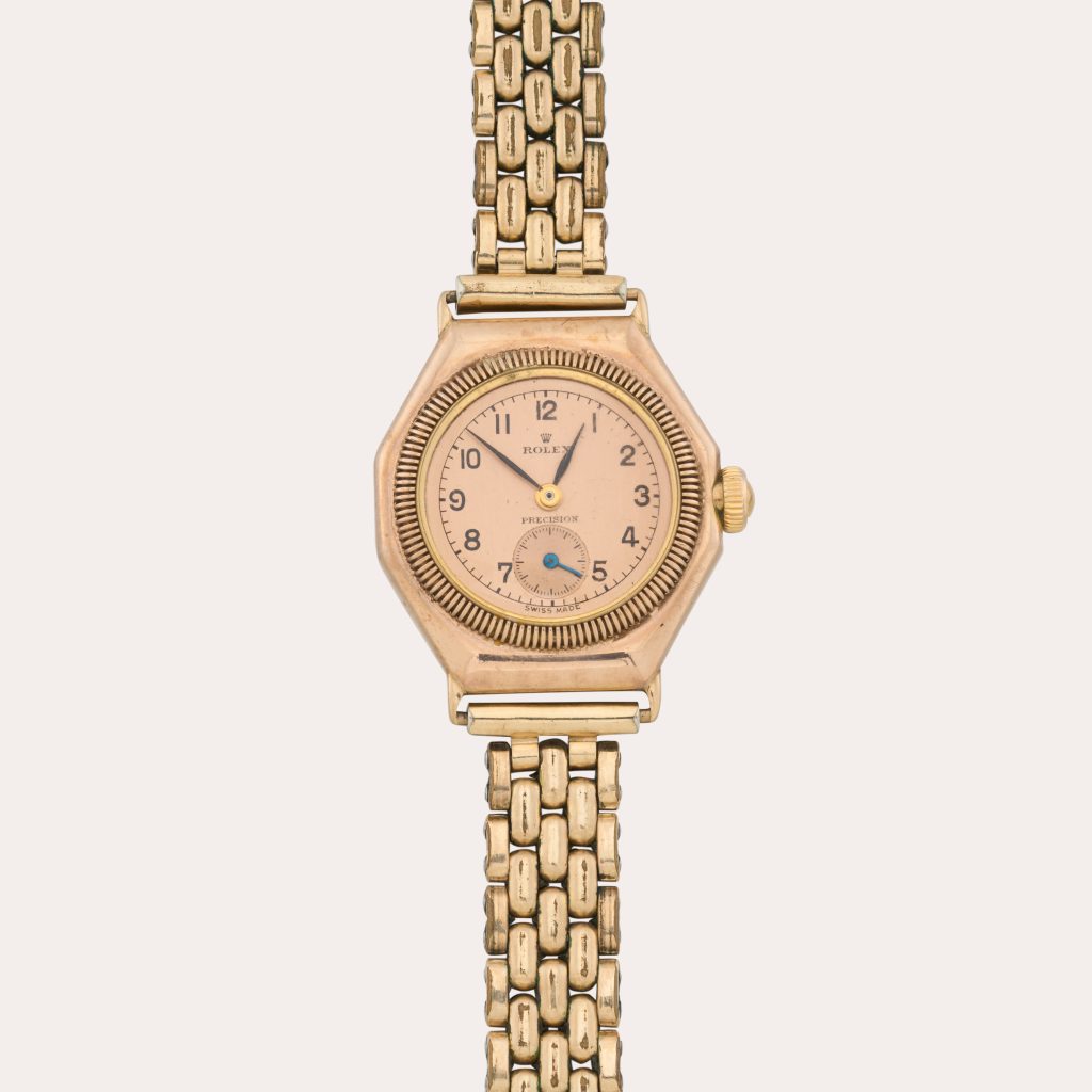 Un orologio da polso Rolex d'epoca in oro, che ricorda lo stile indossato da Mercedes Gleitze, presenta un quadrante rotondo con numeri arabi, un quadrante dei piccoli secondi a ore 6 e un bracciale in oro collegato, su uno sfondo chiaro.