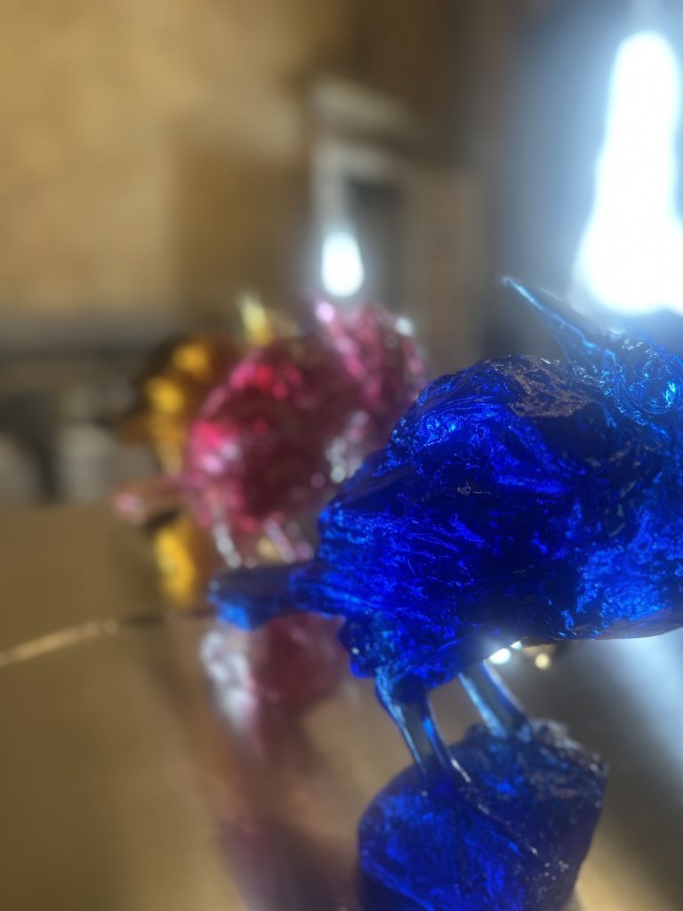 Primo piano di sculture di uccelli colorati, con un uccello blu in primo piano e uccelli dorati e rosa sfocati dietro, su una superficie riflettente. L'influenza del design cinese è evidente nelle forme eleganti e nella lavorazione dettagliata.