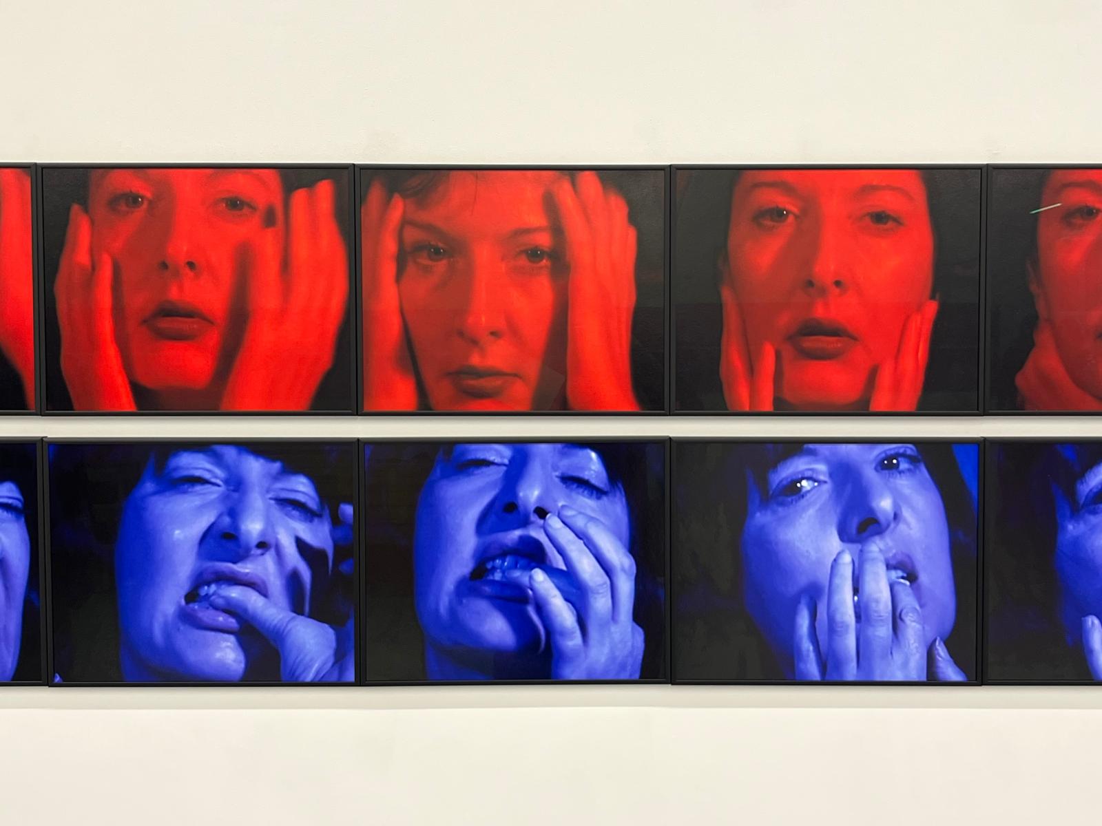 Una serie di otto ritratti fotografici mostra una donna che si tocca il viso - che ricorda Marina Abramović Londra - con la fila superiore in toni rossi e quella inferiore in blu, catturando espressioni facciali intense ed emotive.