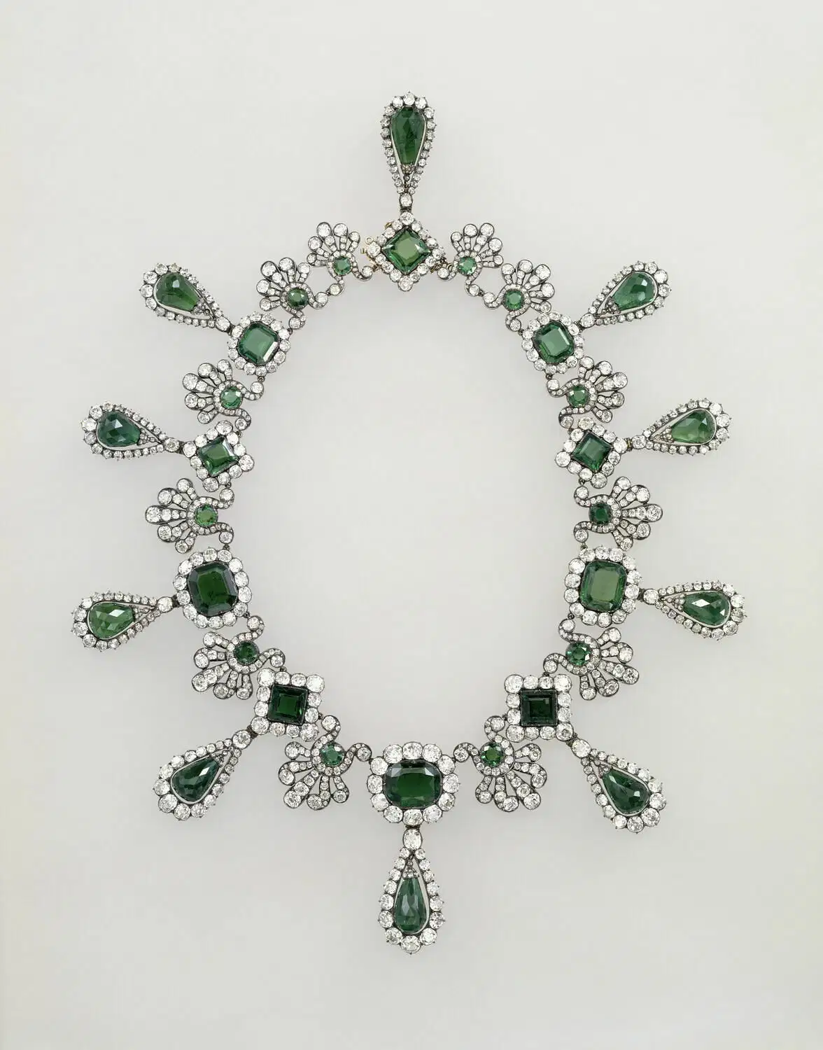 Una collana ornata, che ricorda i tesori del Louvre, presenta gemme verdi di varie forme, circondate da piccole pietre bianche in un motivo floreale simmetrico e a goccia su sfondo bianco.