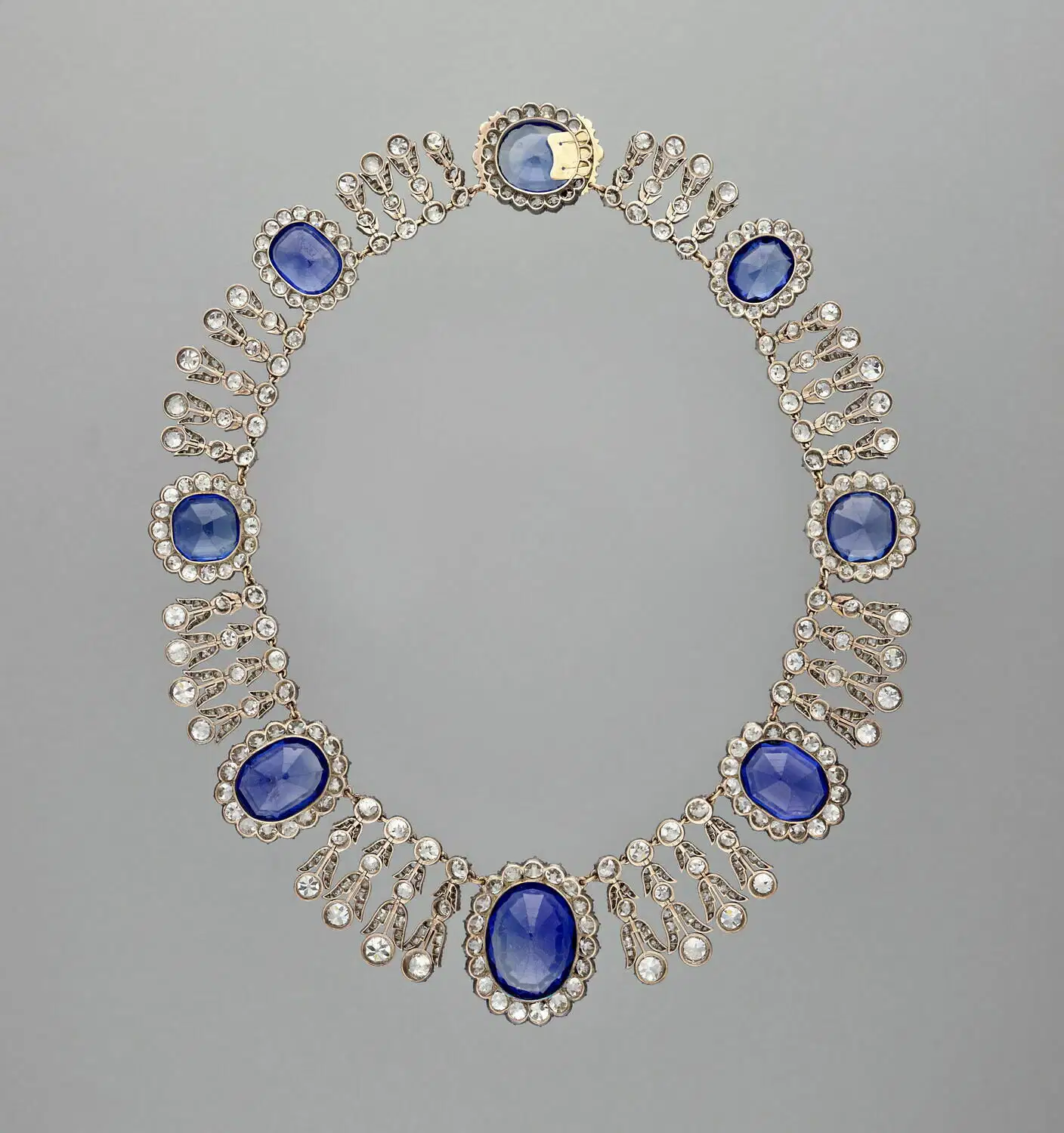 Una collana ispirata al furto gioielli Louvre, con sette grandi gemme ovali blu, ciascuna circondata da piccole pietre chiare, con gruppi di pietre chiare a forma di goccia che si irradiano verso l'esterno, il tutto incastonato in una fascia d'argento su uno sfondo semplice.