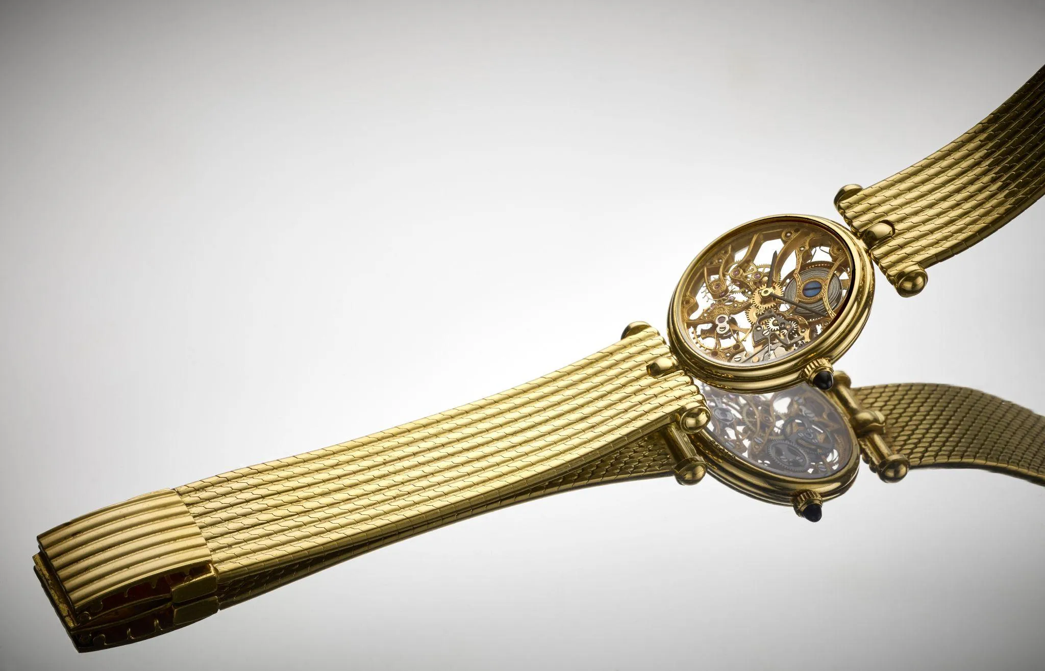 Un orologio da polso scheletrato in oro con cinturino a rete è esposto su una superficie riflettente, che rivela gli intricati ingranaggi e meccanismi all'interno del quadrante rotondo dell'orologio: un vero capolavoro di design orologiero.