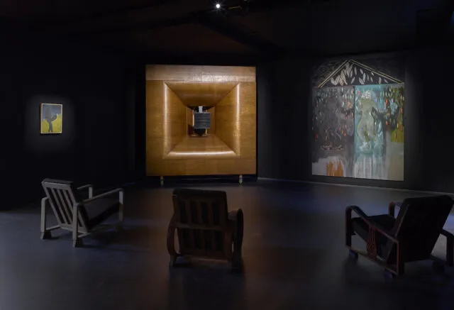Una sala buia della galleria Serpentine, con tre sedie di legno vuote di fronte a una grande installazione geometrica, affiancata da due dipinti su pareti nere, illuminate in modo soffuso da una luce mirata: l'atmosfera di una mostra di Peter Doig.