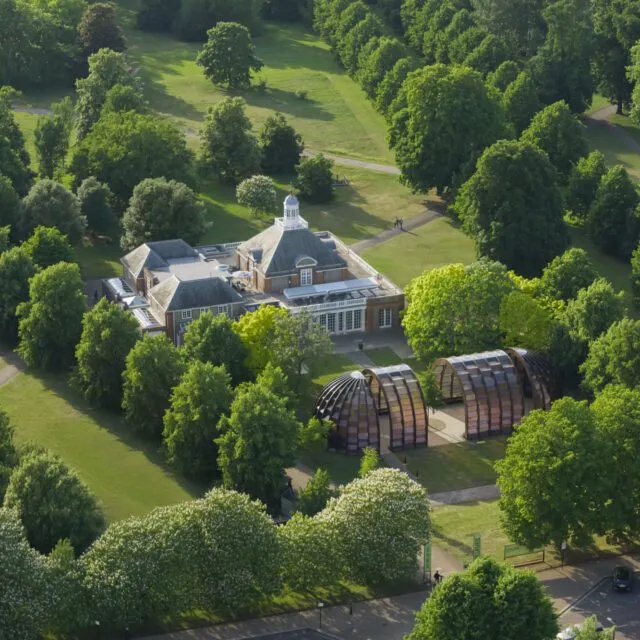 Veduta aerea di un edificio signorile circondato da alberi e prati verdi, con vicino due moderne strutture in legno curvato - che riprendono lo spirito contemporaneo di Peter Doig alla Serpentine - immerso in un grande parco in una giornata di sole.