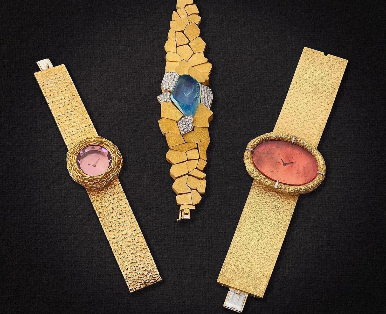 Tre orologi da polso vintage in oro su sfondo nero; ogni segnatempo presenta un design unico: a sinistra il quadrante rosa, a destra il quadrante rosso e al centro l'ampio cinturino in oro con una gemma blu e accenti di diamanti.