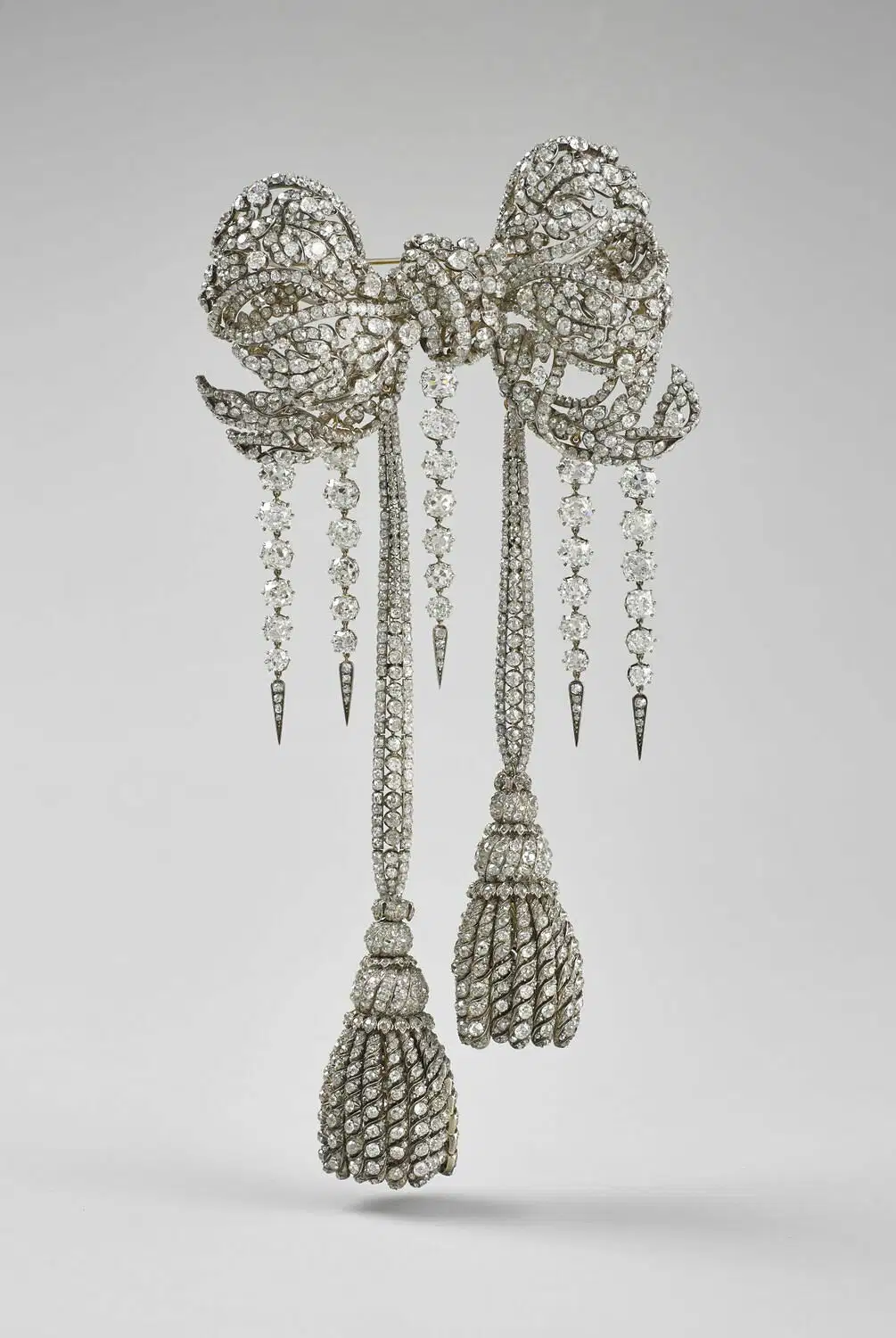 Una spilla ornata di diamanti a forma di fiocco con lunghi pendenti a forma di nappe e catene penzolanti, che ricordano i tesori di cui si parlava nel furto gioielli del Louvre, incastonati in un semplice sfondo grigio.