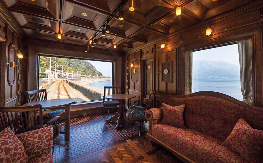 Interni lussuosi di un vagone ferroviario con pannelli in legno, poltrone eleganti, divani e grandi finestre che offrono una vista panoramica della ferrovia costiera, del mare e delle montagne lontane sotto un cielo limpido.
