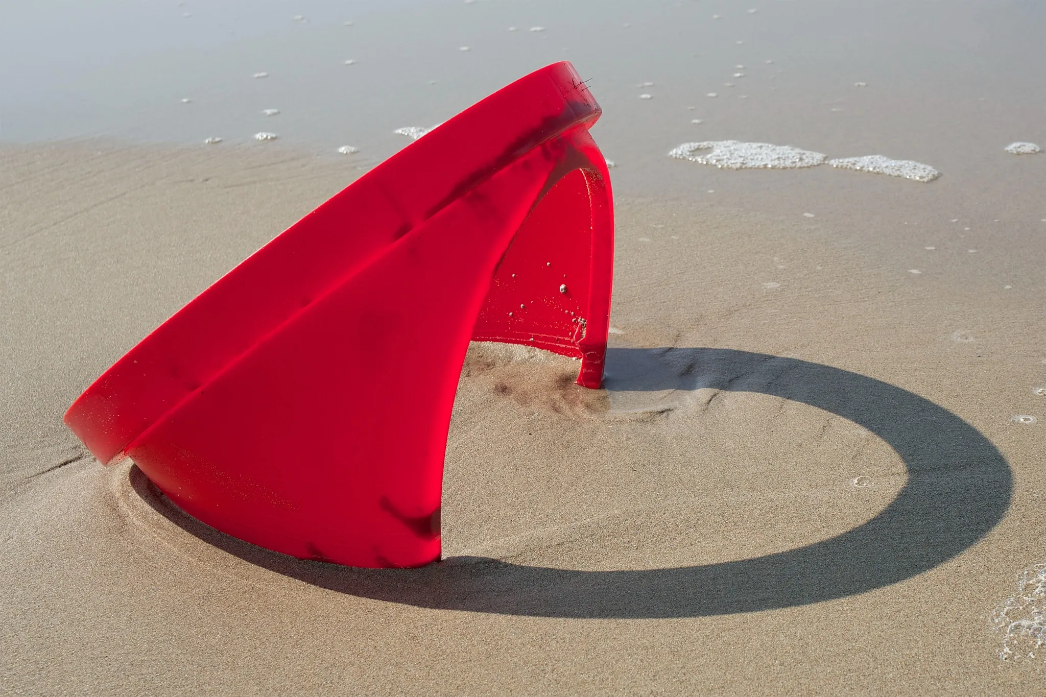 Un secchio di plastica rosso giace su un fianco, parzialmente sepolto nella sabbia bagnata della spiaggia. L'acqua schiumosa del mare è vicina e il secchio proietta un'ombra curva sulla sabbia: una scena che ricorda la suggestiva fotografia di Giuseppe Ripa AIPB.