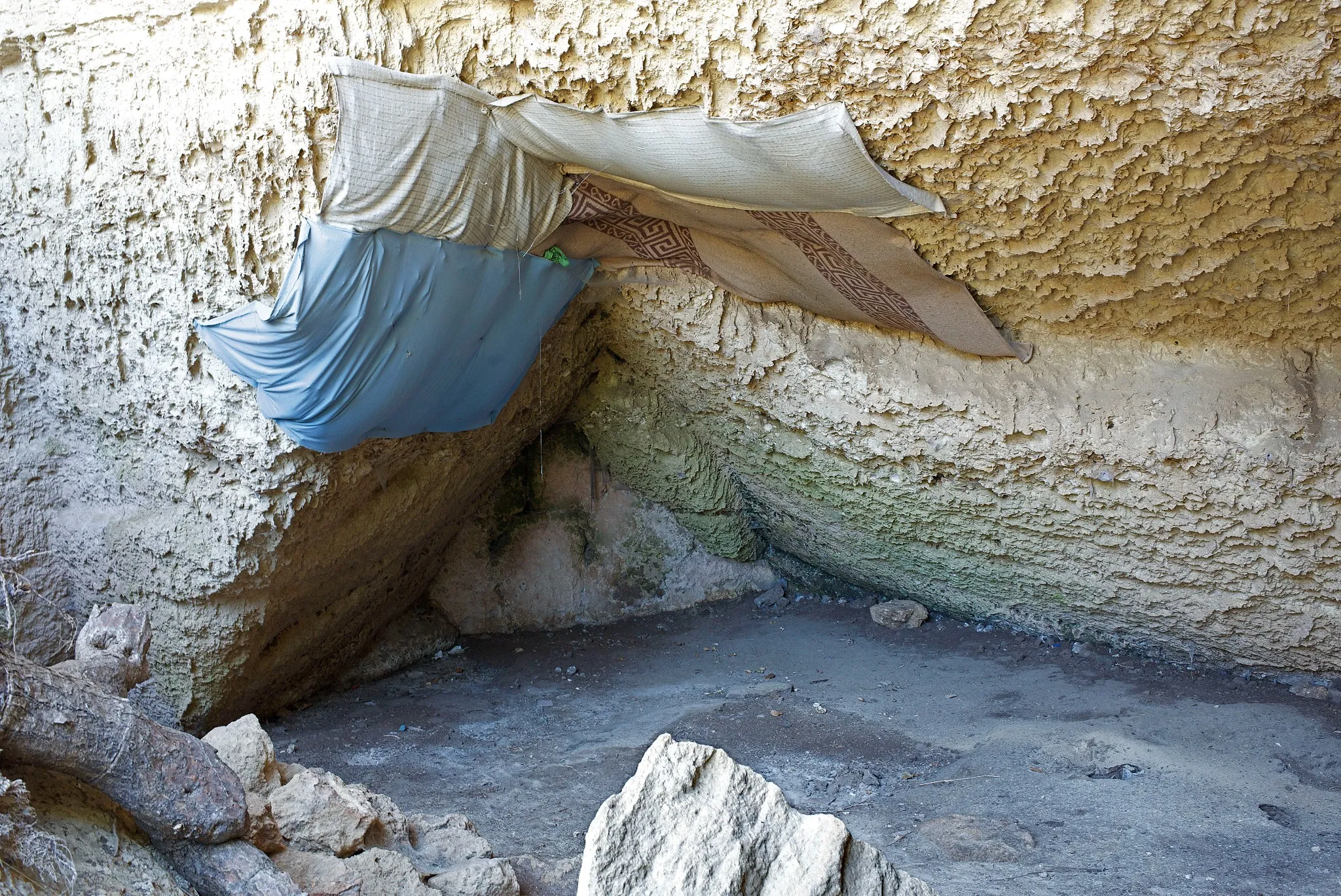 Un riparo di fortuna con coperte e lenzuola drappeggiate su un'area rocciosa, simile a una grotta, che crea uno spazio ombreggiato contro le pareti e il pavimento in pietra - una scena suggestiva immortalata da Giuseppe Ripa AIPB - con rocce sparse in primo piano.