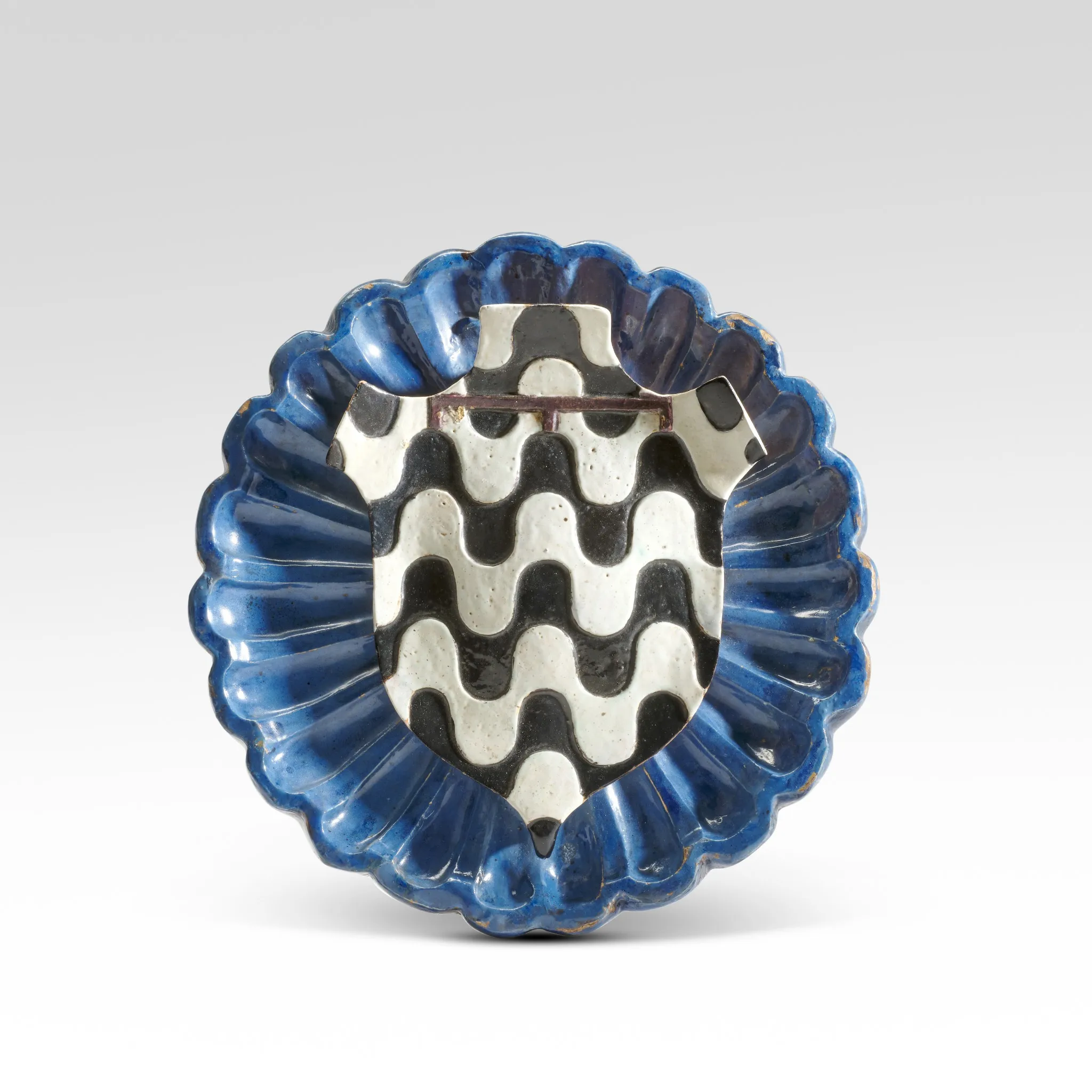 Un piatto in ceramica smerlato con un bordo blu e uno scudo centrale in argento con un motivo ondulato in bianco e nero, questo pezzo vanta una superficie lucida e riflettente - il suo design simmetrico ricorda i precedenti record di Sotheby's Italia.
