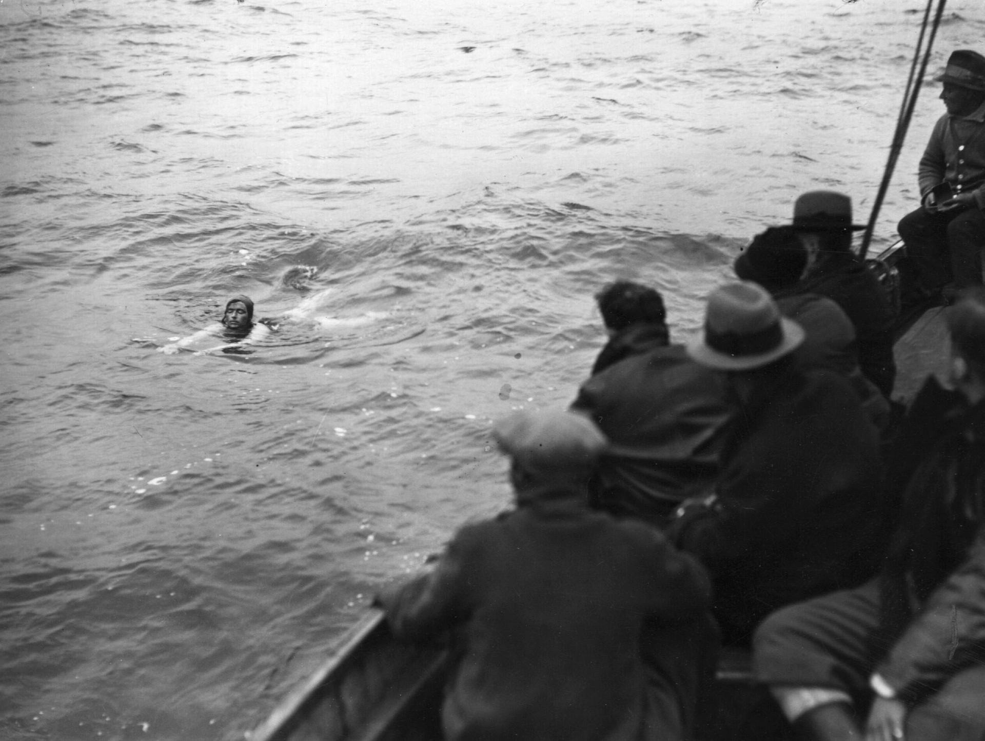 Una nuotatrice, che ricorda le famose imprese di resistenza di Mercedes Gleitze con il suo fidato Rolex, lotta contro l'acqua agitata mentre diverse persone in cappotto e cappello osservano con tensione da una barca vicina. Le condizioni difficili accentuano la drammaticità della scena.