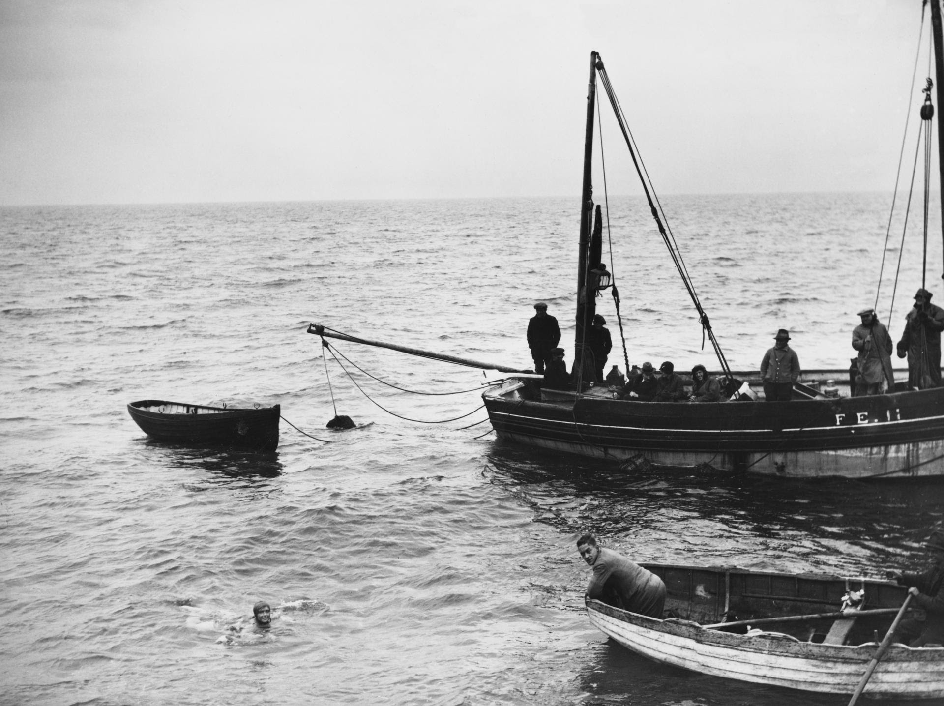 Una foto in bianco e nero mostra persone su due piccole barche di legno in mare. Una persona, che ricorda la nuotata Rolex di Mercedes Gleitze, è in acqua vicino alle barche, mentre diverse altre osservano dalle barche sotto un cielo nuvoloso.