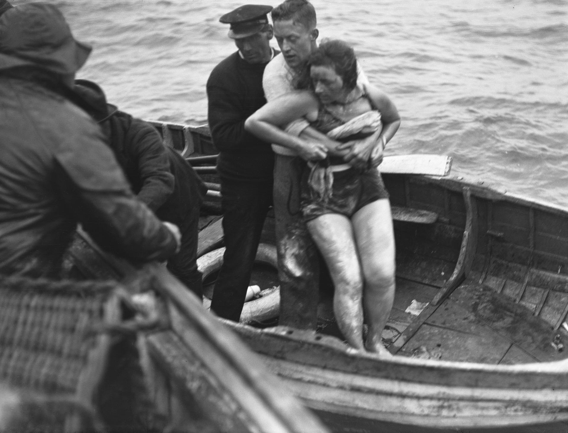 Un uomo aiuta una donna in costume da bagno, che ricorda Mercedes Gleitze, che indossava notoriamente un Rolex, a salire su una barca di legno. Altri uomini assistono mentre intorno a loro si vedono onde e acque agitate.