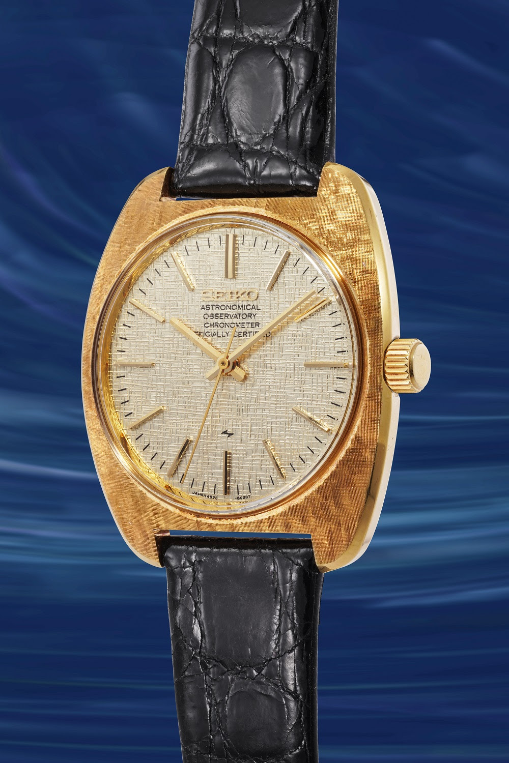 Un orologio da polso d'oro con quadrante chiaro, lancette d'oro sottili e cinturino in pelle nera - come gli orologi giapponesi lusso - si staglia su uno sfondo blu e vorticoso. Il quadrante dell'orologio presenta un piccolo testo con indicazioni dettagliate.