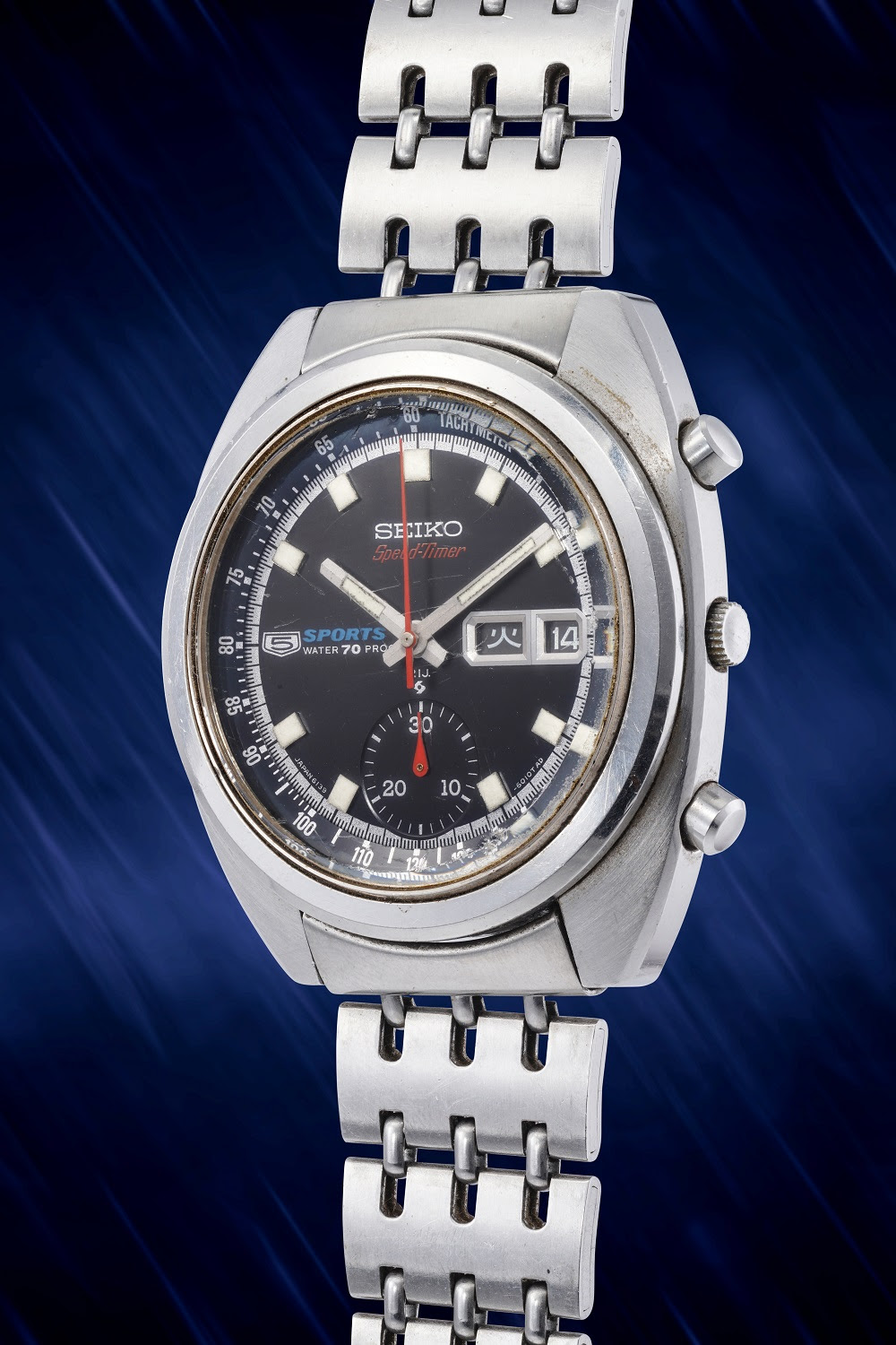 Un orologio da polso Seiko Sports 5 Speedtimer d'epoca, che incarna orologi giapponesi di lusso, con cinturino in metallo argentato e quadrante nero, è esposto ad angolo su un suggestivo sfondo blu striato: un reperto ideale in qualsiasi asta.