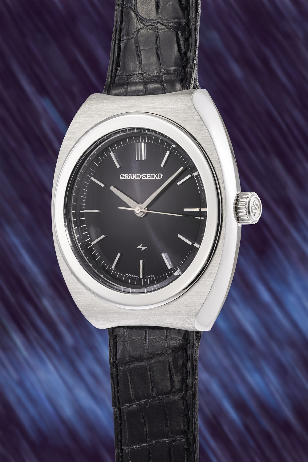 Un orologio da polso Grand Seiko color argento con cinturino in pelle nera e quadrante nero, esposto in posizione verticale su uno sfondo a strisce blu e viola: uno squisito esempio di orologi giapponesi di lusso pronto per qualsiasi asta esclusiva.