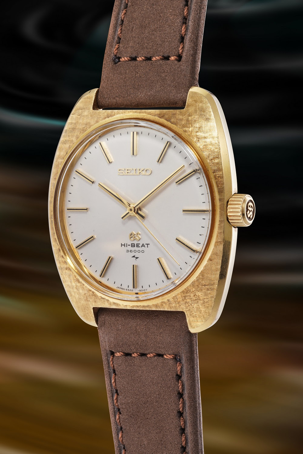 Un orologio da polso Seiko in oro con quadrante bianco, indici e lancette in oro e cinturino in pelle marrone, su uno sfondo multicolore sfocato: uno squisito esempio di orologi giapponesi di lusso offerti all'asta. Il quadrante riporta la dicitura "HI-BEAT 36000" al di sopra delle ore 6.