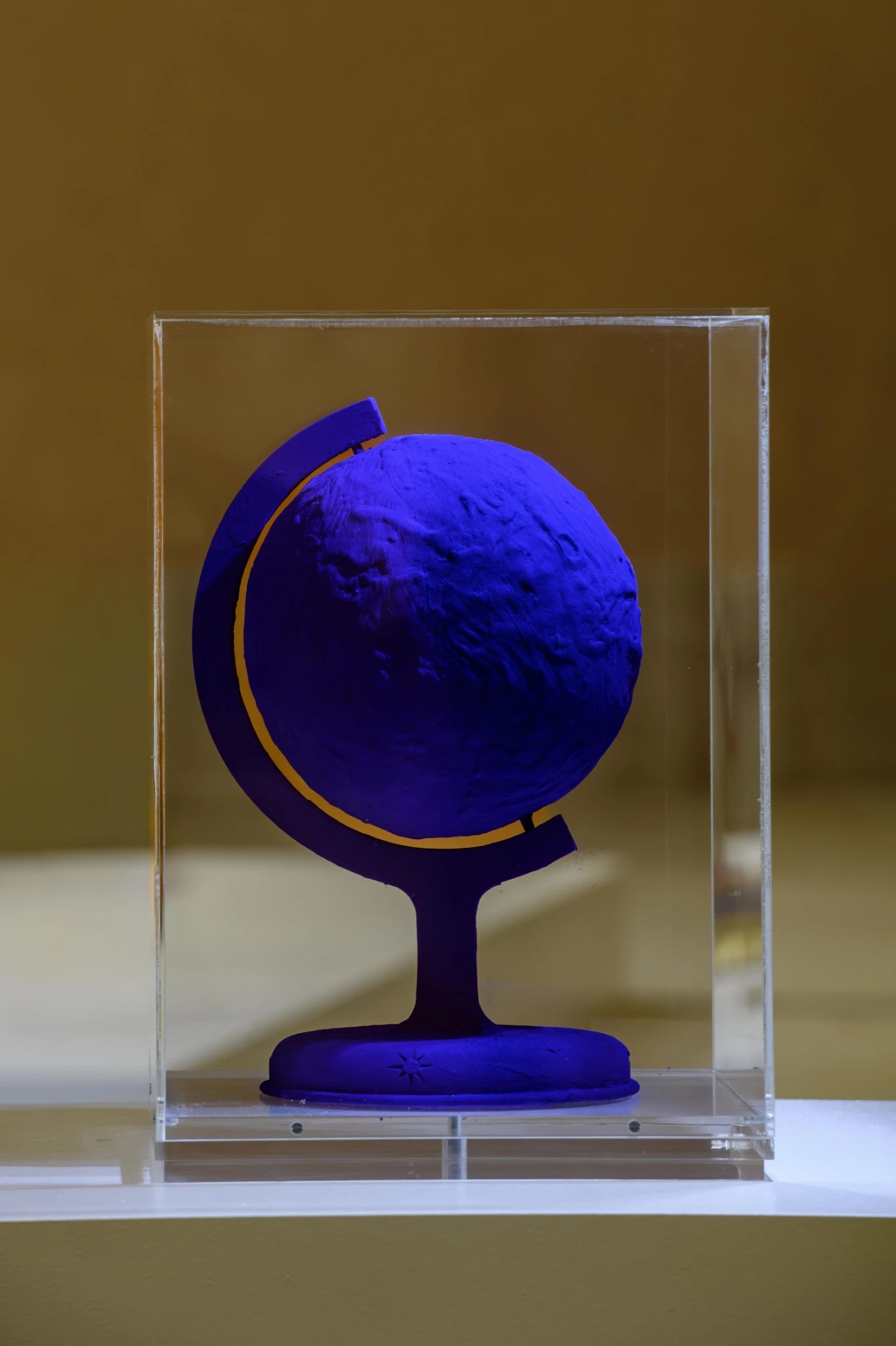 Una scultura di un mappamondo dipinto in blu Yves Klein con un meridiano dorato, esposto all'interno di una teca in vetro o acrilico trasparente su uno sfondo beige.