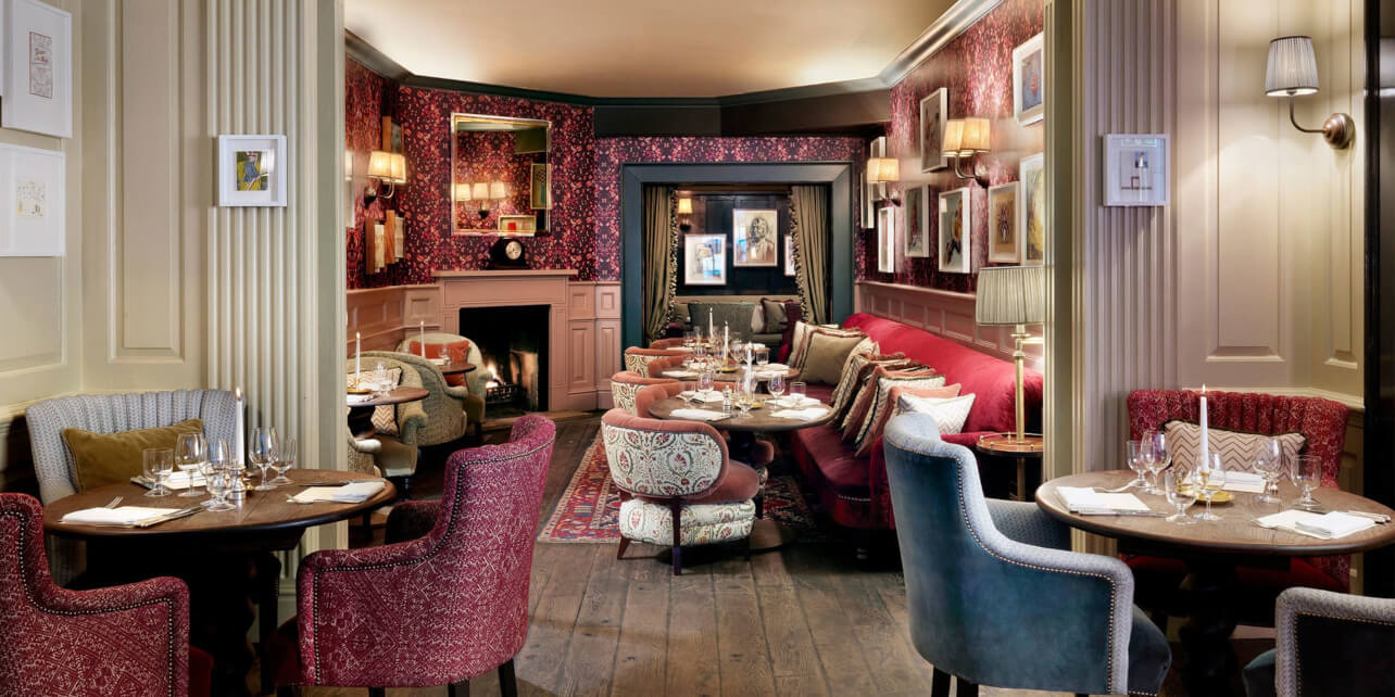 A cozy, elegant restaurant with patterned armchairs, red and blue seating, dark wood floors, and framed art on red-patterned walls. Tables are set for dining, and a fireplace adds warmth to the inviting atmosphere.