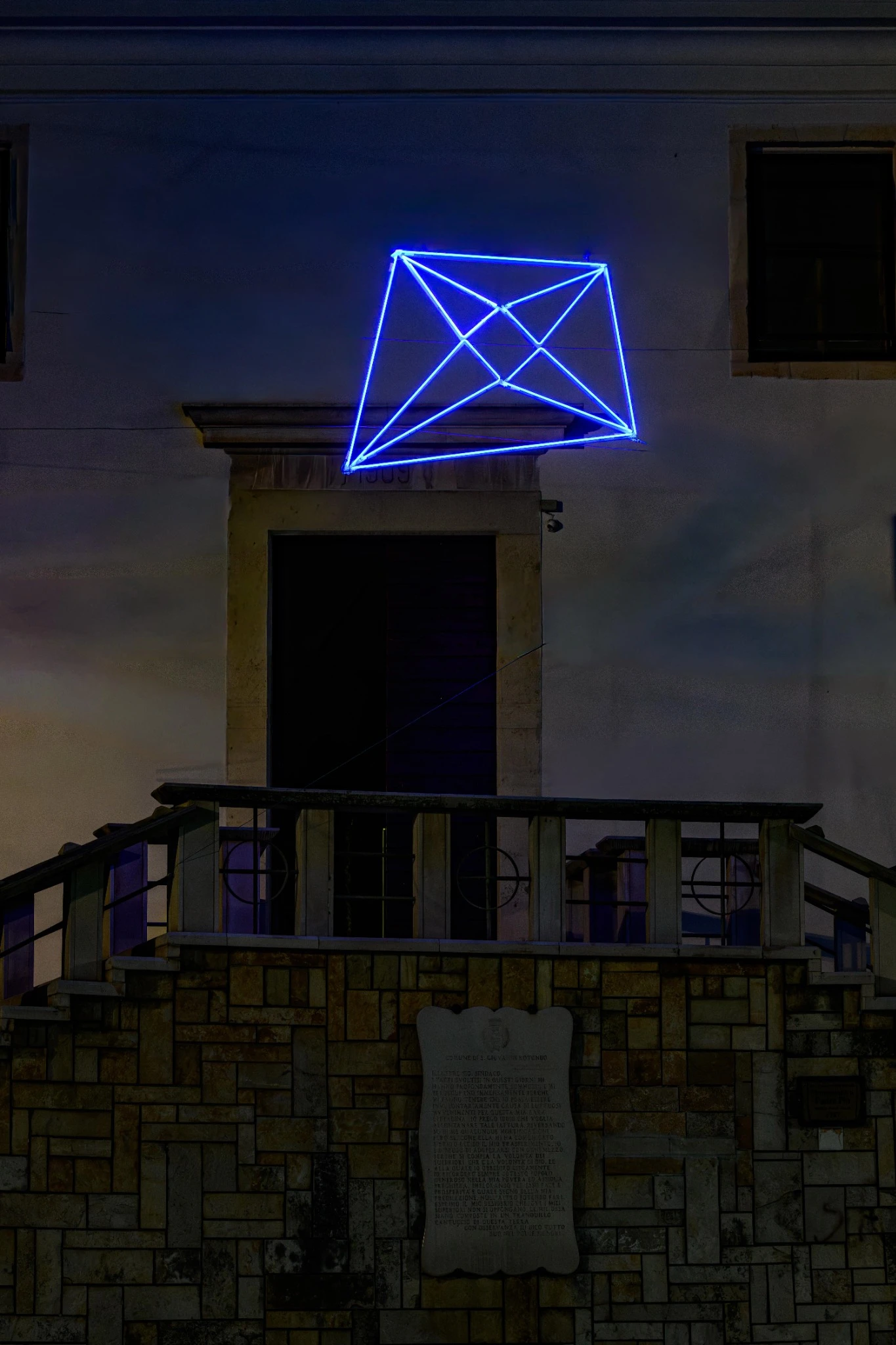 Un'insegna al neon a forma di aquilone Yves Klein Blu è montata di notte sopra una porta di un edificio in pietra, con una targa e una ringhiera in pietra visibili in primo piano.