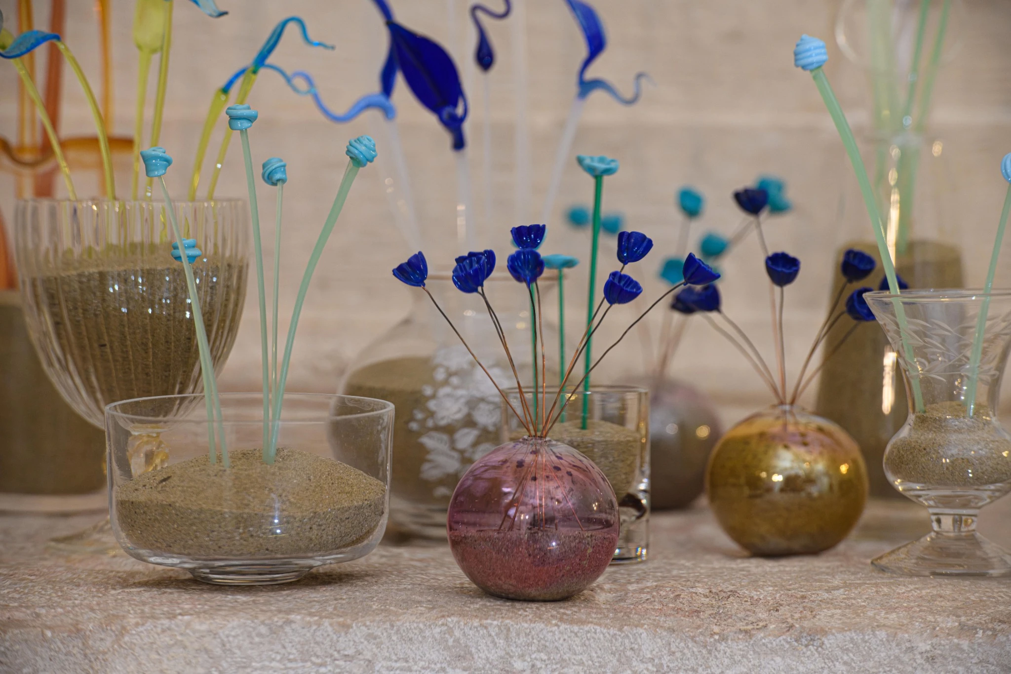 Vasi decorativi in vetro riempiti di sabbia e steli colorati in vetro soffiato con forme di fiori blu e turchesi di Yves Klein, disposti su una superficie di pietra su uno sfondo neutro.