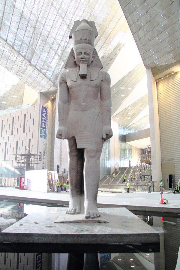Una grande statua egizia di un faraone si trova nell'ampio atrio del nuovo museo egizio, con l'alto soffitto di vetro che inonda l'area di luce mentre sullo sfondo si muovono alcuni operai in giubbotto di sicurezza.