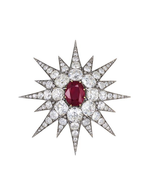 Una spilla a forma di stella con un rubino ovale centrale circondato da punte a raggiera con diamanti bianchi rotondi e diamanti blu mellon, che creano un design scintillante e simmetrico.