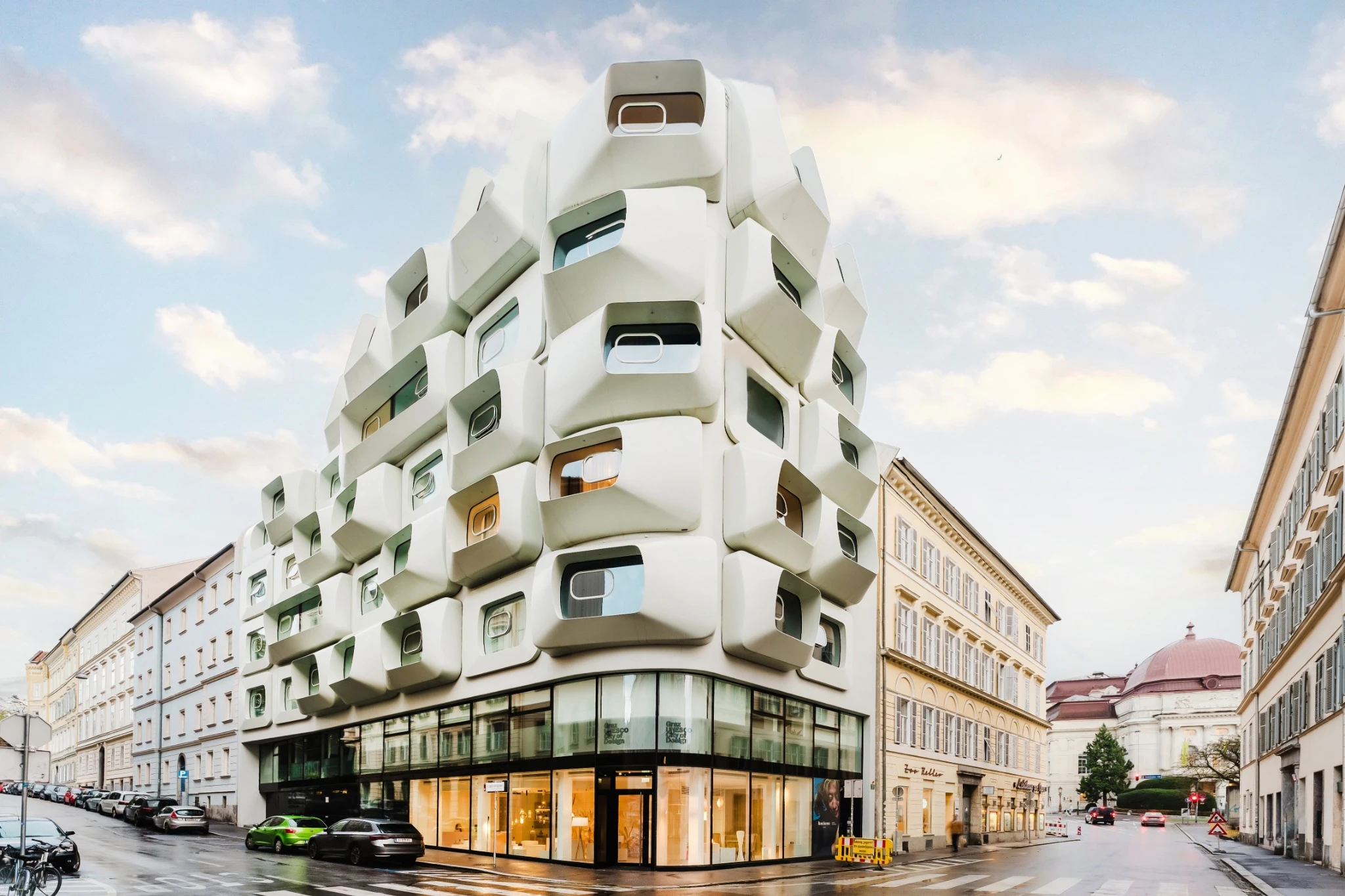 Un edificio moderno con una singolare facciata geometrica bianca con cornici di finestre sporgenti si trova all'angolo di una strada, mescolando Abu Dhabi lusso con gli edifici tradizionali beige circostanti in un'area urbana sotto un cielo nuvoloso.