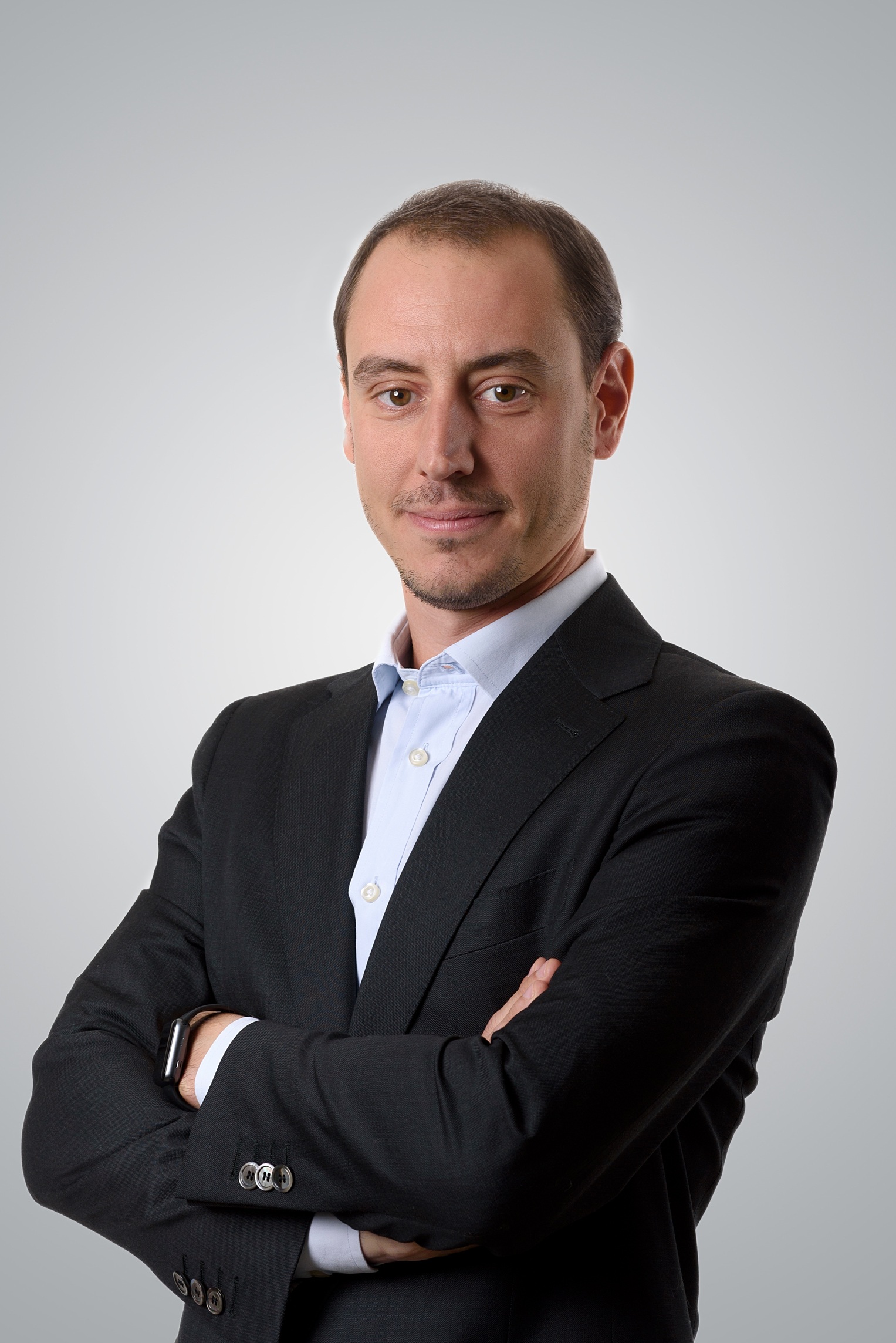 Un uomo in abito scuro e camicia bianca sta in piedi con le braccia incrociate, guardando con sicurezza la telecamera su uno sfondo grigio chiaro, incarnando l'essenza di un'arte patrimoniale alternativa per le immagini professionali contemporanee.