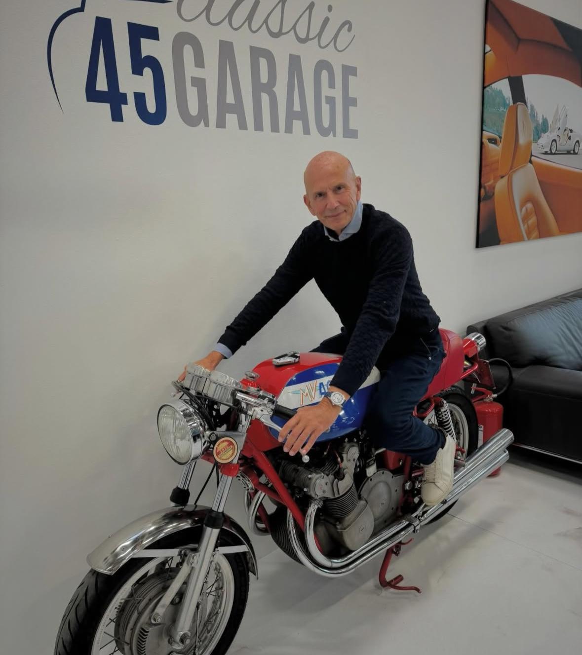 Un uomo con capelli corti e grigi e abbigliamento casual siede su un'automobile d'epoca in un moderno garage con pareti bianche e decorazioni a tema automobilistico. La scritta "Classic 45 GARAGE" è visibile sulla parete alle sue spalle.