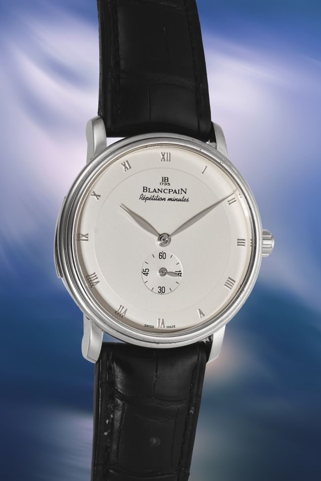 Un orologio da polso di lusso Blancpain che ricorda un orologio Coppola, con quadrante rotondo argentato, indici a numeri romani, quadrante dei piccoli secondi a ore 6 e cinturino in pelle nera su uno sfondo sfumato blu e viola.