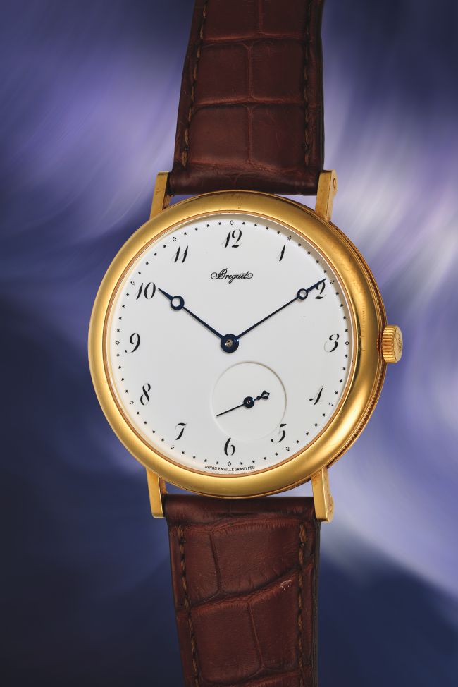 Classico orologio da polso Coppola Orologio in oro con quadrante bianco, lancette blu, numeri arabi, quadrantino dei piccoli secondi e cinturino in pelle marrone, su uno sfondo sfocato viola e bianco.