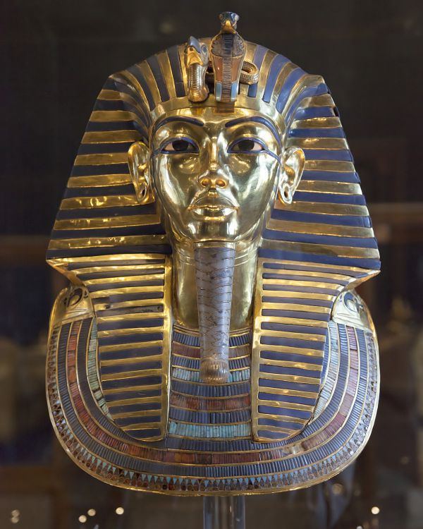 L'immagine mostra la maschera funeraria dorata di Tutankhamon, con un copricapo a strisce, dettagli intarsiati in blu e oro e un cobra e un avvoltoio sulla fronte, esposta nel nuovo museo egizio su uno sfondo scuro.