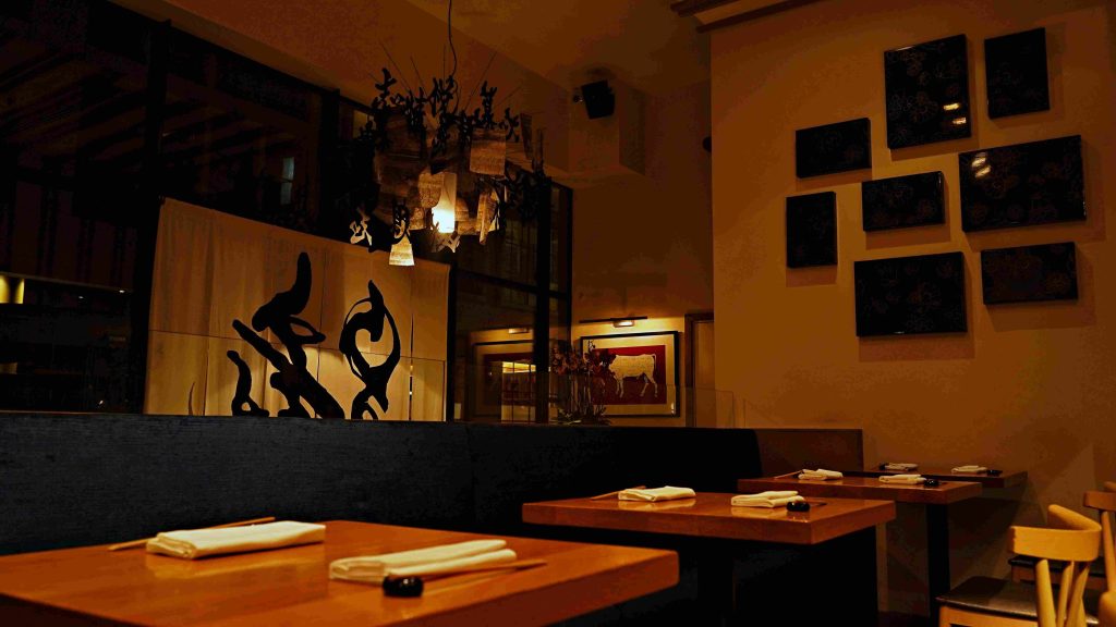 A dimly lit restaurant with wooden tables set with napkins and chopsticks, dark bench seating, a decorative divider with abstract designs, and framed art on cream-colored walls.