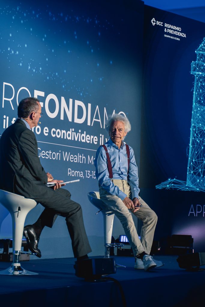 Due uomini siedono sul palco per discutere di una conferenza. Uno indossa un abito, l'altro una camicia azzurra con bretelle. Dietro di loro c'è un fondale blu con il testo di un evento e l'illustrazione digitale di un faro.