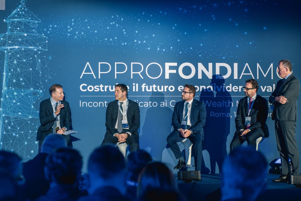 Cinque uomini in abito da lavoro siedono e parlano su un palco davanti a un fondale blu con testo in italiano e una grafica di rete digitale, suggerendo una tavola rotonda formale a una conferenza o a un evento professionale.