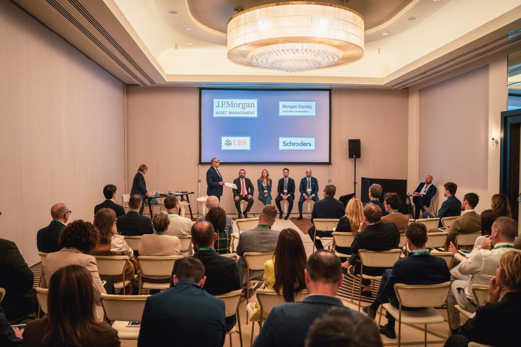Un gruppo di persone partecipa a una tavola rotonda commerciale in una sala conferenze. Cinque oratori siedono sul palco, mentre il pubblico ascolta. I loghi di JP Morgan, Morgan Stanley, UBS e Schroders sono visualizzati sugli schermi alle loro spalle.