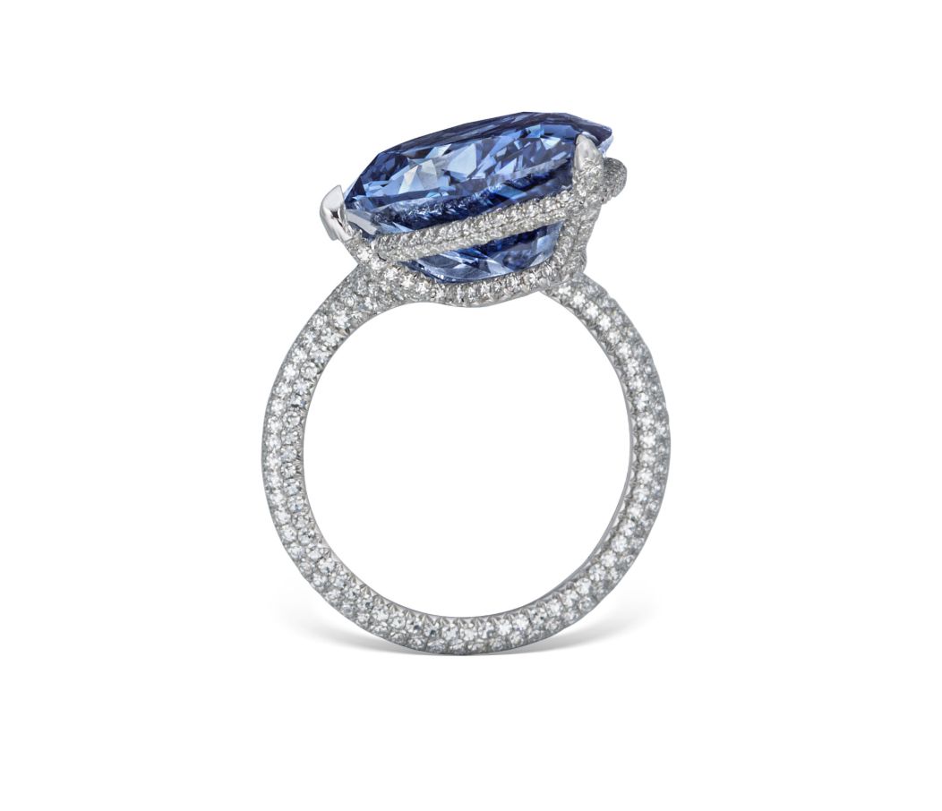 Lussuoso anello con diamante blu mellon, caratterizzato da una grande gemma di forma ovale incastonata in una fascia tempestata di piccoli diamanti bianchi, su sfondo bianco.