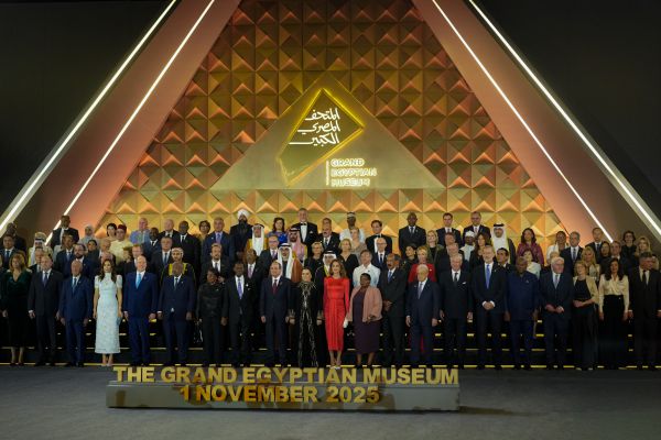 Un folto gruppo di persone vestite in modo formale si trova sul palco davanti a un fondale triangolare dorato con le scritte "The Grand Egyptian Museum", "nuovo museo egizio" e "1 novembre 2025".